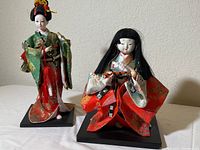 Lot includes one standing Japanese Geisha figure dressed in green kimono with gold and red details and one seated figure with long black hair in red and white kimono, both on black bases.