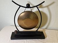 Full view of brass dinner gong mounted on black metal curved stand and wooden base with mallet resting on base.