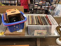 Blue basket with 45 RPM records and clear plastic bin filled with vinyl albums and 45s on a table, showing the quantity and storage containers.