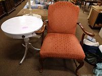 Photo of a white painted round Mersman accent table with pan feet next to a vintage orange upholstered wooden armchair showing the overall lot condition and detailing.