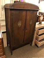 Front view of wooden armoire showing double doors with floral decorations, curved top, and castors below.