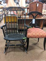 Two chairs side by side: a black Bombay Company exotic chair with upholstered red and gold floral seat and decorative back; next to a black painted Hitchcock-style rocking chair with gold accents.