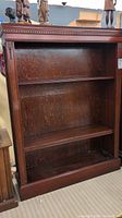Front view of dark mahogany finish wood bookcase showing three adjustable shelves and carved decorative details along top and sides.