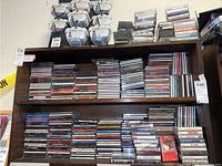 Shelf with multiple stacked CDs and some cassette tapes on top, showing many titles and cases.