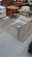 Overall view of the square glass coffee table showing metal ornate legs and clear glass top on a carpet.