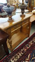 Front and side view of wood console table showing drawer, turned legs, and bottom shelf with two decorative lamps on top.