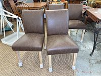 Four brown leather-like dining chairs with straight backs and padded seats. Light wooden legs with white floor protectors visible. Chairs arranged in two front and two back positions.