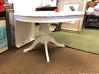 Full view of the round white wooden dining table showing the tabletop and pedestal base with signs of wear and paint chipping.