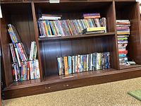 Wide view of a wooden shelving unit containing numerous DVDs organized across three compartments. Titles include TV series, children's movies, and general movies. Cases vary in color and style, some stacked vertically, others horizontally.