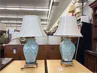 Two matching ceramic turquoise lamps with diamond pattern and gold-tone bases topped with white bell-shaped fabric shades, displayed on wooden surfaces in a showroom.