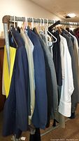 Side view of several men's clothing items on hangers on a pipe rack including blazers, jackets and shirts in mixed colors such as navy, black, grey, white, and beige.