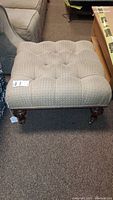 Front image of large square ottoman with tufted beige fabric upholstery, wood carved legs, and brass caster wheels.
