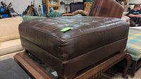 Large, dark brown, oversized leather-like ottoman with button tufting visible on the top surface. Shows general wear and creasing on the upholstery.