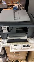 Front view of HP OfficeJet Pro 9025 printer with front panel open showing ink cartridge slots, paper output tray, and some cords around it.