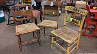 Photo showing three vintage wooden chairs with painted backrests and natural rush seats, arranged together, displaying their overall appearance and condition.