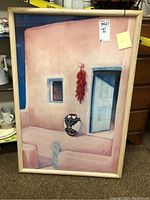 Framed signed painting showing a Mexican style home front with pink stucco, blue door, chili peppers, and pot, with stain on top of frame.