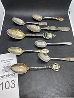 Top-down photo of eight sterling silver souvenir spoons arranged on a black surface showing varied designs and enamel details.