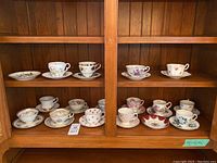 Wide shot of 15 teacups each paired with a saucer plus 2 extra saucers displayed in wooden cabinet.