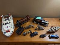 Overview photo of all transportation toys on a wooden surface showing the white plastic boat, brown metal train and many metal motorcycles and cars.