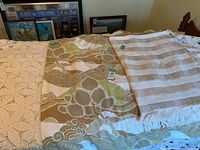 Photo showing three vintage bed coverings: a large white hand-crocheted bedspread, a patterned fabric cover with floral and abstract design, and a beige and white checkerboard woven throw with fringe.