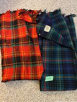 Two folded vintage wool plaid blankets side by side on carpet; one red-orange plaid, one green-blue plaid with label.