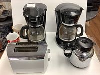 Photo showing two black Sunbeam coffee makers with glass carafes, thermal carafe, two mugs, and a white Proctor Silex toaster on a table.