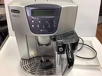 Front view of the silver espresso maker showing the control panel with buttons and display, drip tray and attached milk frother container with handle.