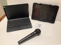 Zagg keyboard iPad case open with iPad, Survivor rugged case with iPad, and black wireless microphone on table