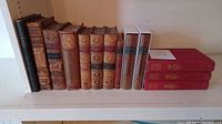 Wide shot showing all 13 books on a shelf, including antique leather-bound books, two contemporary books in original sleeves, and three red hardcovers.