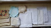 Top view of assorted crocheted doilies, pastel blue napkins, and folded white Irish linen tablecloths and napkins on a surface.