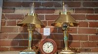 Pair of brass oil style table lamps with glass chimneys and conical brass shades flanking a Pottery Barn clock with wooden frame and Roman numeral face against a brick wall.