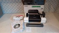 Overview of Canon Pixma iP110 printer kit in original box with power adapter, power cord, ink cartridges, manuals, and installation discs laid out on surface.