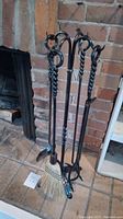 Full view of the black wrought iron fireplace tool set including stand, broom, poker, tongs, and shovel against brick background.