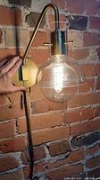 Right side view of aged brass/gold wall sconce attached to rough brick wall with clear glass shade and pull chain switch.