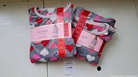 Two pairs of new unopened Kate Spade two-piece pajamas in size Medium with heart pattern on gray fabric.