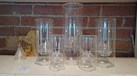 Full set of five cylindrical glass oil lamps with varying heights and accessories on shelf against brick wall background