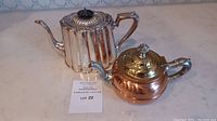 Photo showing both the silver plate ribbed teapot and the copper teapot on a marble surface, with auction label visible.