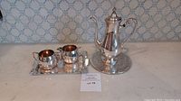 Complete silver plated tea set on counter, includes teapot or coffee pot, creamer, sugar bowl on a tray, with a separate round trivet.