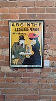 Full view of the framed vintage Absinthe poster hanging on a brick wall, showing the man and woman at a table with Absinthe bottles.