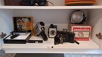 Wide shelf view displaying several vintage cameras and accessories including Kodak Instamatic M24 in original box, Cameralite box, flash units and folding cameras.