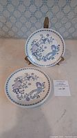 Two plates shown, one upright on a stand, displaying the blue and purple mid-century modern figures and floral pattern.