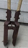 Close-up image of the metal blades and wooden handles of a manual post hole digger, showing rust and wear on the metal parts.