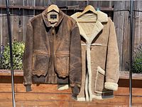 Two men's jackets hanging from wooden hangers on a metal rack outdoors. Left jacket is brown distressed leather bomber by Scully with collar, zip front, ribbed cuffs and two flap pockets. Right jacket is tan shearling coat lined with cream shearling visible at collar, lapels, cuffs, and pockets.