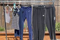 Six pieces of Nike activewear hung on a clothing rack outdoors, including tops and bottoms in blue, black, and striped patterns.
