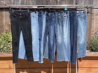 Nine pairs of designer denim jeans hanging on a rack outdoors in various shades of blue and black, showcasing different washes and styles.