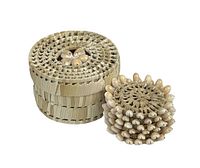 Woven raffia straw cylindrical canister with a lid decorated by puka shells arranged in a flower pattern. Canister is closed and shows natural beige coloration and intricate weaving.