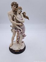 Full view of the painted statue depicting a nude male figure embracing a woman in a pink dress, mounted on a round wooden base.