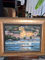 Frontal view of framed oil painting showing coastal harbor scene with boat and shoreline buildings