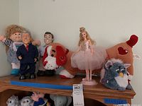 Shelf display of dolls including Sugar Plum Barbie, Clinton doll, Cabbage Patch Kid, Furby, and plush dog toy.