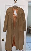Full view of camel hair cashmere coat hanging on a door showing the front, interior satin lining is visible, camel color, size tag attached